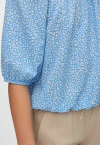 Lichtblauwe blouse met korte, opgeblazen mouwen die een luipaardprintpatroon in het wit heeft. De stof lijkt textuur te hebben, met geplooide accenten aan de onderrand.