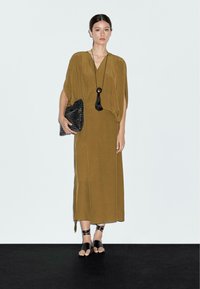 Robe maxi vert olive avec des manches amples, présentant des détails de couture verticaux. Le modèle tient une pochette texturée noire et porte des sandales noires à lanières.