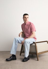 Chemise à carreaux rouge et blanc à manches courtes, jeans droits bleu clair, et mocassins en cuir noir. Assis sur une chaise à revêtement clair.
