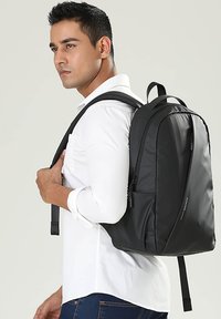 Sac à dos noir avec une finition lisse et mate, doté de bretelles ergonomiques, de poches zippées et d'un design rectangulaire mince.