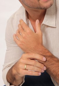 Auksinė vestuvinė žiedas ant vidutinės rudos odos rankos, dėvinčios šviesios spalvos lininę marškinių dalį. Rankos atpalaiduotos, matomi smulkūs detalės ir tekstūra.