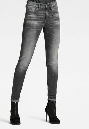 Žena má na sobě vybledlé černé úzké džíny s vyhrnutými lemy a černé kotníkové boty na vysokém podpatku se zipy vpředu, stojí před jednobarevným pozadím.