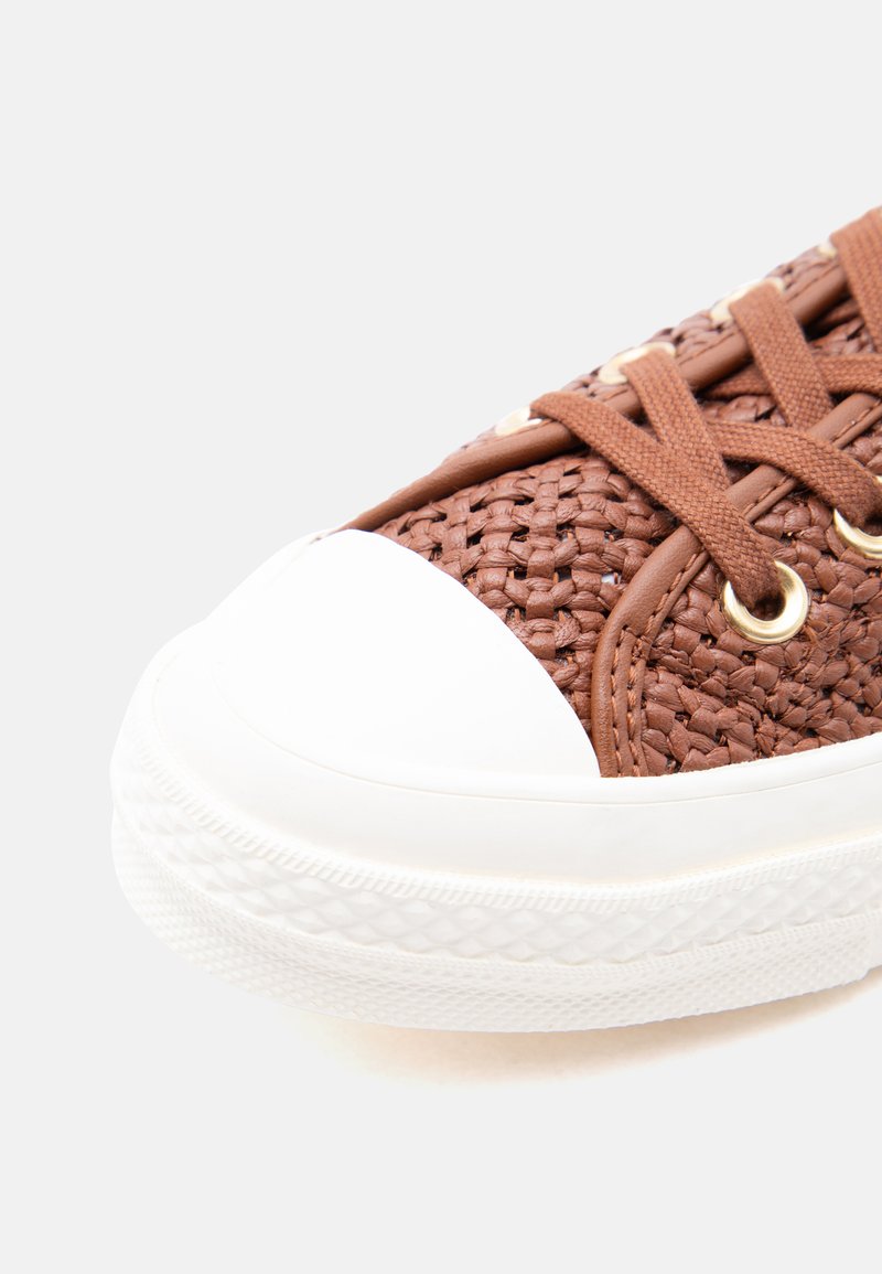 Brown woven sneaker with white rubber toe cap and sole, featuring brown laces threaded through gold eyelets on a white background.