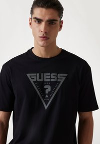 Juodas medvilninis marškinėliai su dideliu pilku trikampiu grafiku, kuriame yra tekstas "GUESS", klausimo ženklas ir mažesnis tekstas žemiau. Trumpomis rankovėmis.