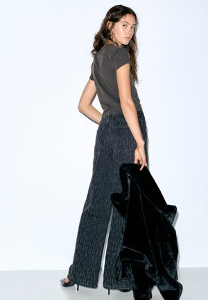 Siv bombažna majica, široke črne hlače z teksturo in črna šala iz umetnega krzna. Model stoji z obrnjenimi hrbtom, obut v sandale s paščki.