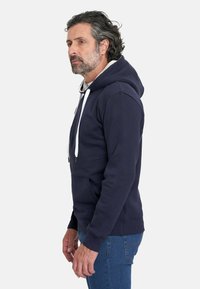 Sudadera azul marino hecha de tela suave, cuenta con capucha ajustable, bolsillo tipo canguro y puños acanalados. Modelo mostrado de perfil.