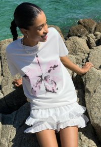 T-shirt blanc avec un imprimé floral rose, associé à une jupe blanche à volants. Modèle posé sur des rochers près de l'eau.