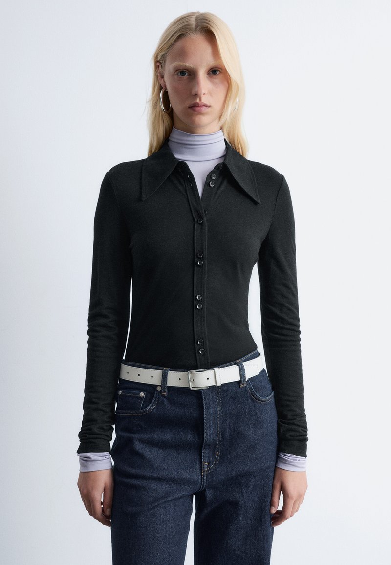 Black long-sleeve shirt with a pointed collar, buttoned down front, layered over a light purple turtleneck, paired with blue jeans and a white belt.