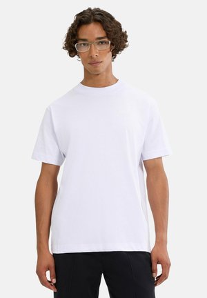 Junger Mann mit lockigem Haar und Brille, der ein schlichtes weißes T-Shirt und schwarze Hosen vor hellem Hintergrund trägt.