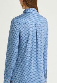 Camicia blu a fantasia con colletto, caratterizzata da un disegno geometrico in un blu più scuro. Maniche lunghe con dettagli a bottoni e piega posteriore.