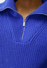 Pull-over bleu tricoté avec un col haut et une fermeture à demi-fermeture éclair. Tissu texturé avec un motif côtelé et un décolleté en V prononcé.