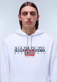 Sudadera blanca con un gráfico impreso en azul marino y rojo, que muestra el nombre de la marca "NAPAPIJRI", con un diseño de bandera y cierre con cordón.