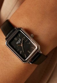 Rechthoekig zwart horloge met zilveren accenten, zwarte leren band en minimalistische wijzerplaat met zilveren uurmarkeringen en merklogo.