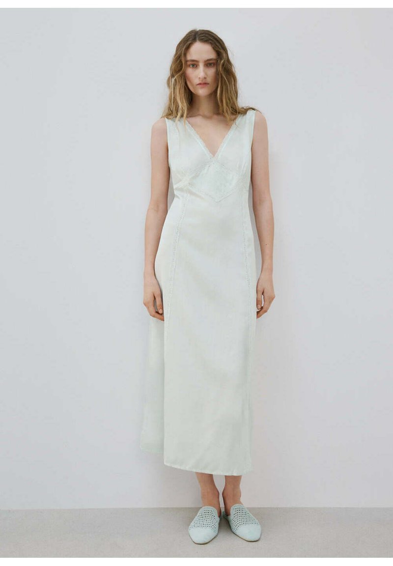 Femme aux longs cheveux ondulés portant une robe midi blanche sans manches avec un col en V et des chaussures blanches tissées faciles à enfiler, debout contre un mur uni.