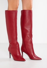 Bottes en cuir rouge jusqu'au genou avec une finition texturée, bout pointu et talon aiguille fin. Design lisse et ajusté sans quincaillerie visible.