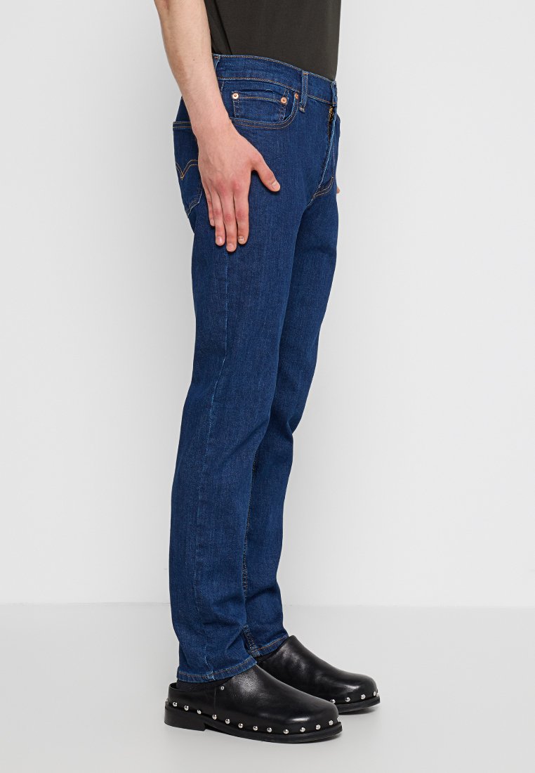 Denim jeans in dark blue, straight fit design with orange stitching and five pockets. Paired with black clogs featuring metal studs.