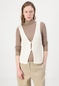 Weißer, gerippter Strickwesten mit tiefem V-Ausschnitt und einer vorderen Öffnung, getragen über einem braunen Rollkragenpullover. Die Weste hat eine figurbetonte Form und eine strukturierte Oberfläche.