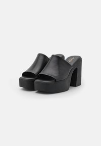 Svarte platåhæler laget av glatt skinn, med åpen tå og bred stroppdesign, samt chunky høye hæler og teksturert såle.