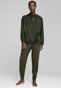 Olivgrüner Kapuzenpullover mit Kängurutasche, kombiniert mit dazu passenden, schmal zulaufenden Hosen, die ein kleines Logo auf der linken Hüfte tragen.