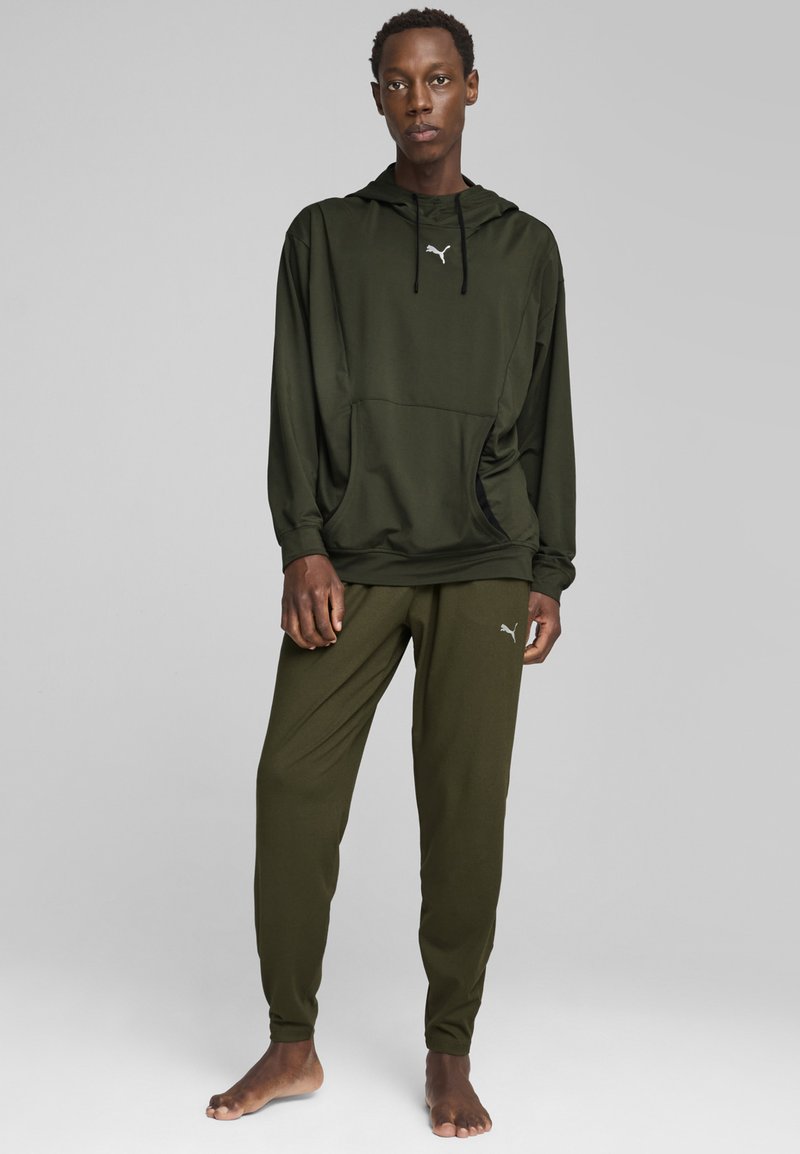 Olivgrüner Kapuzenpullover mit Kängurutasche, kombiniert mit dazu passenden, schmal zulaufenden Hosen, die ein kleines Logo auf der linken Hüfte tragen.