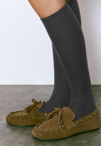 Mocasines de ante marrón con lazo anudado y acentos de ojales metálicos, combinados con calcetas de algodón gris de altura hasta la rodilla. Diseño de punta redonda y plana.