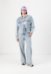 Chaqueta de mezclilla de color azul claro con bolsillos frontales, combinada con unos jeans de pierna ancha a juego. Completado con una gorra lavanda con un diseño de rosa.