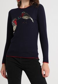 Maglione blu navy con bordo rosso con due uccelli di paillettes rivolti l'uno verso l'altro, abbinato a pantaloni neri.