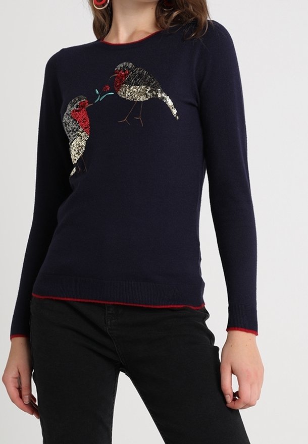 Maglione blu navy con bordo rosso con due uccelli di paillettes rivolti l'uno verso l'altro, abbinato a pantaloni neri.