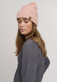 Strikket rosa lue med et teksturert mønster og en dusk på toppen, med et lite brunt logomerke. Enkelt og varmt design.