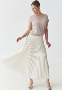 Blusa beige chiaro con scollo a V abbinata a una lunga gonna bianca plissettata. La gonna presenta pieghe verticali e una silhouette fluida. Indossata con sandali con punta aperta.
