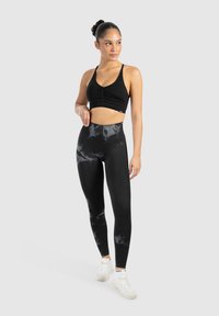 Donna in piedi con una mano sui fianchi, che indossa un reggiseno sportivo nero, leggings high-waisted neri con motivo tie-dye, calze bianche e scarpe da ginnastica bianche su sfondo grigio.