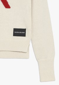 Světle krémový pletený svetr s červeným designem, žebrovanými manžetami a lemem, a černou etiketou s nápisem "Calvin Klein Jeans" v bílé barvě.