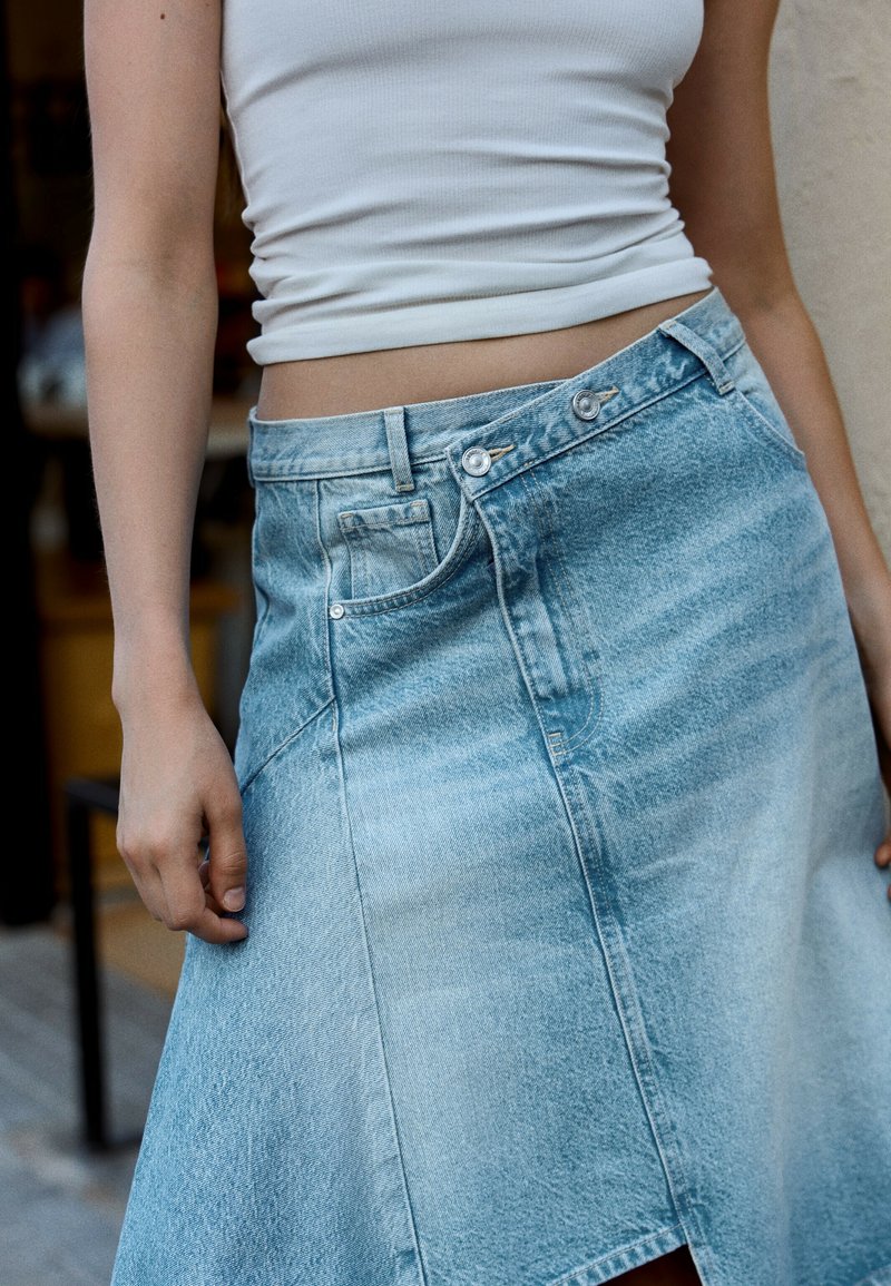 Personne portant un débardeur court blanc et une jupe en denim bleu avec une ceinture à double bouton superposée.