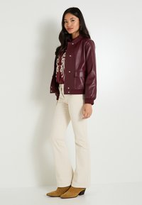 Mujer de pie con pantalones acampanados beige, botas de tacón color marrón, chaqueta de cuero burdeos y camisa de patrón contra un fondo blanco liso.