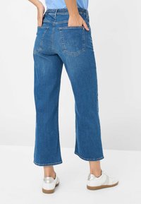 Blauwe denim wijde broek met een hoge tailleband, twee achterzakken en een onafgewerkte zoom, gecombineerd met witte sneakers met zilveren accenten.