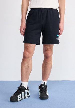 Mand iført sorte Nike-shorts med blåt logo og sorte Nike Air-sneakers, stående på blåt gulv med hvide sokker.