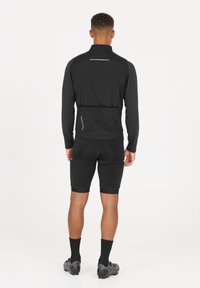 Schwarze Herren-Radjacke mit langen Ärmeln, reflektierenden Akzenten und taillierter Passform über schwarzen Shorts und Fahrradschoenen mit strukturierten Sohlen.