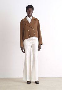 Veste courte brune à double boutonnage avec col texturé, chemise blanche à boutons et pantalon blanc évasé. Chaussures noires à bouts pointus.