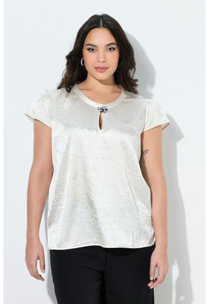 Kvinde med mørkt hår iført en cremefarvet satinbluse med korte ærmer, nøglehul foran og dekorativ spænde, kombineret med sorte bukser.