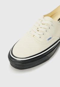 Canvas sneaker in off-white with black rubber sole. Features white laces, a branded tag, and a black stripe along the sole edge.
