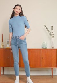Blusa de gola alta em azul claro com textura, combinada com jeans skinny azuis claros. Modelo a usar sapatilhas brancas, de pé contra um fundo neutro.