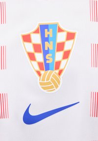 Εμβλημα ποδοσφαίρου της Κροατίας με κόκκινο και λευκό καρό, μπλε κάθετη λωρίδα με τα γράμματα HNS και μια χρυσή μπάλα ποδοσφαίρου, πάνω από μπλε λογότυπο Nike σε λευκό ύφασμα.
