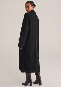 Sieviete ar tumšiem matiem, sapītu zirgasti, ģērbusies garā melnā adītā mētelī un tumši brūnās augstpapēžu kurpēs, pozē pret neitrālu fona.