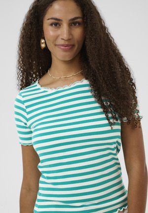 Femeie cu păr lung și creț, purtând cercei de tip cerc aurii, un colier auriu și o cămașă cu mâneci scurte cu dungi albe și verzi.