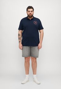 Polo cu culoare bleumarin, cu logo rotund roșu, mâneci scurte, asortat cu pantaloni scurți din denim gri și adidași albi. Materialul este neted, de ajustare lejeră.