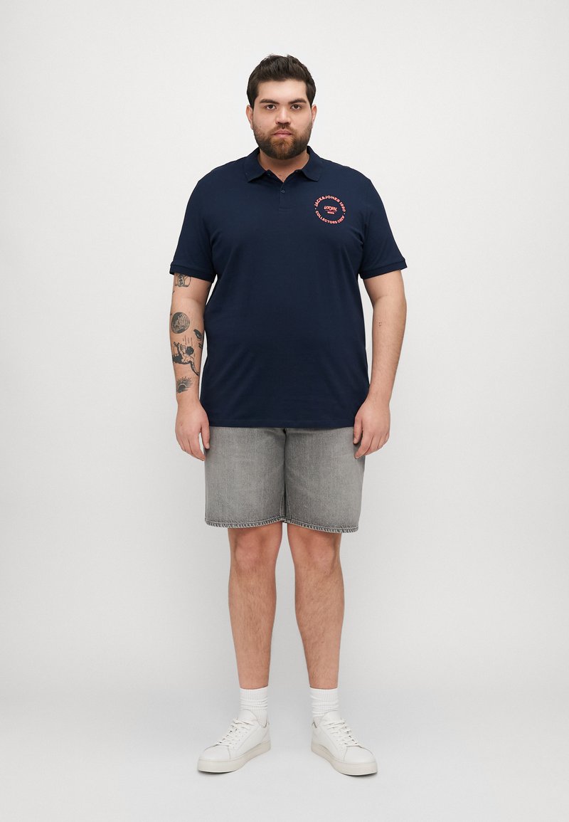 Polo cu culoare bleumarin, cu logo rotund roșu, mâneci scurte, asortat cu pantaloni scurți din denim gri și adidași albi. Materialul este neted, de ajustare lejeră.