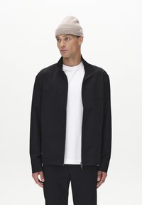 Veste zippée noire en tissu lisse avec col montant, associée à un t-shirt blanc et un bonnet gris clair. Design simple et minimaliste.