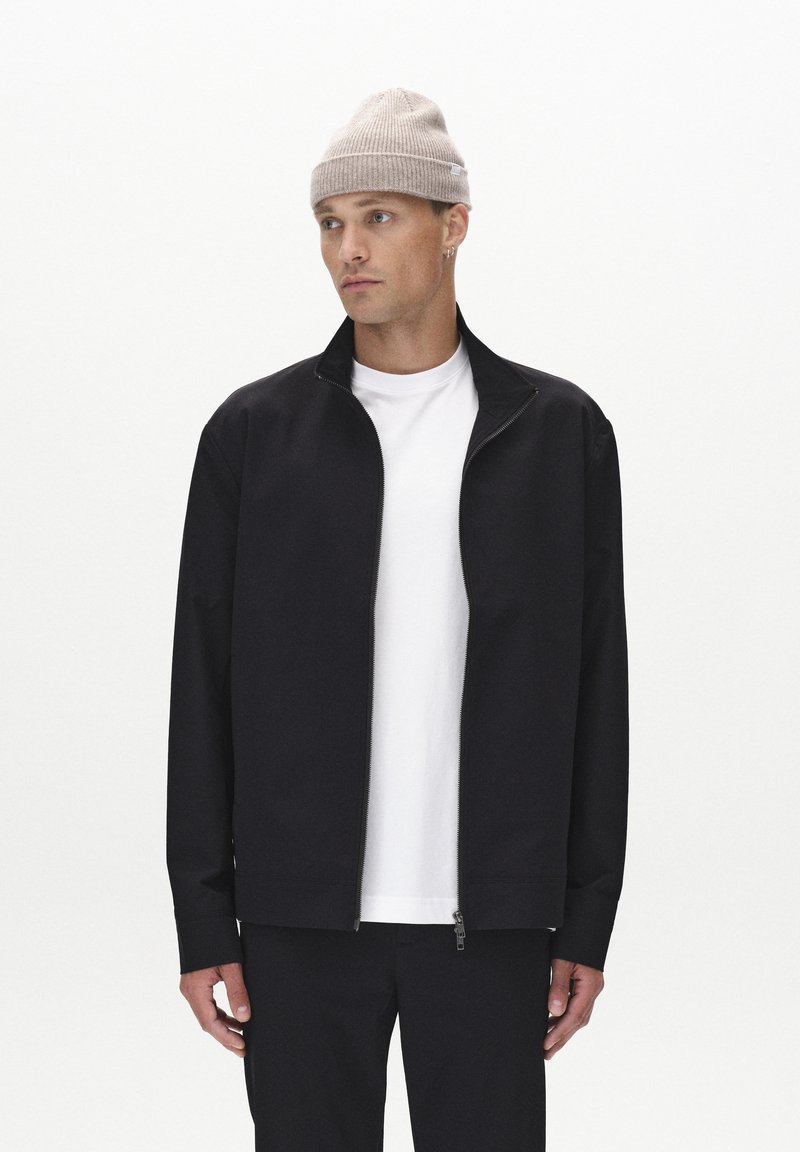 Veste zippée noire en tissu lisse avec col montant, associée à un t-shirt blanc et un bonnet gris clair. Design simple et minimaliste.