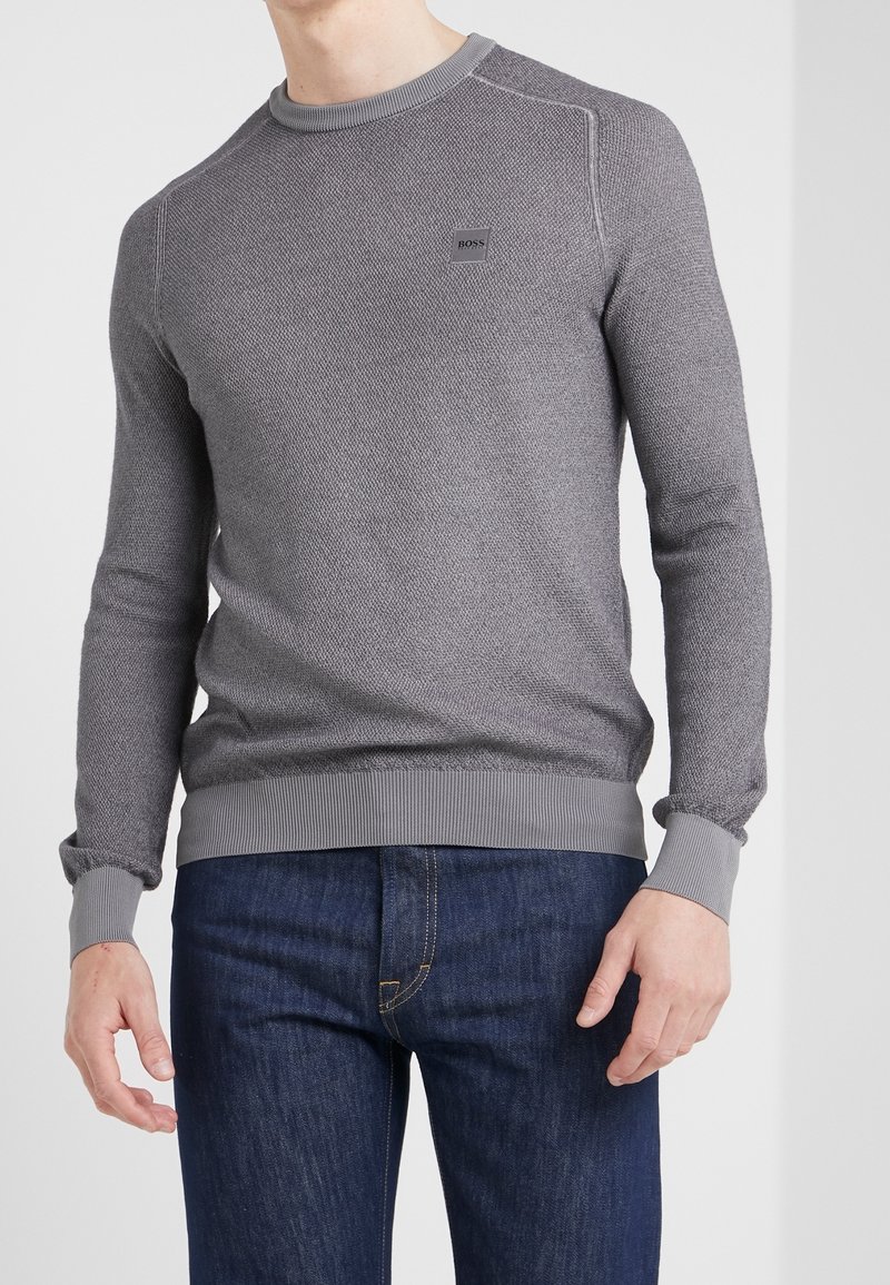 Homme portant un pull gris ajusté BOSS avec un tricot texturé et des poignets côtelés, associé à un jean bleu foncé, debout devant un fond uni.