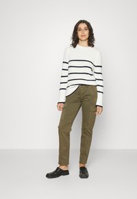 Suéter de malha branco e riscas azul-marinho combinado com calças cargo verde-oliva. As sapatos são tipo slip-on pretos; as calças têm bolsos laterais.